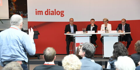 Vier Podiumsteilnehmer stehen an einer Theke und schauen ins Publikum. Im Publikum steht ein Mann am Mikrofon und stellt eine Frage ans Podium.