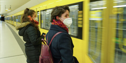 Reisende stehen mit FFP2-Maske an einem U-Bahnhof und warten auf die einfahrende Bahn.