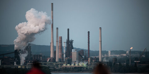 Das Stahlwerk von Thyssenkrupp in Duisburg ist von der Halde Rheinpreussen zu sehen, während Schiffe über den Rhein fahren und zwei Personen im Vordergrund sitzen.