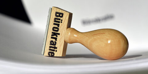 Ein Stempel mit der Aufschrift „Bürokratie“ liegt auf Papieren.