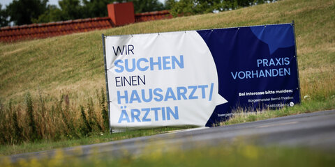 Ein Plakat mit dem Text "Wir suchen einen Hausarzt / Ärztin – Praxis Vorhanden" steht an einem Ortseingang.