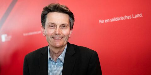 Rolf Mützenich steht vor einer roten Pressewand und schaut lächelnd in die Kamera