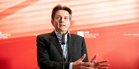 Rolf Mützenich steht vor einer roten Wand und spricht in ein Mikrofon vor ihm.