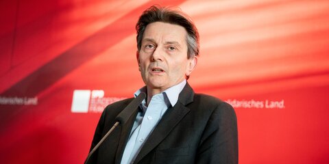 Rolf Mützenich steht vor einer roten Wand und spricht in ein Mikrofon vor ihm.