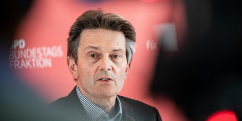 Rolf Mützenich steht vor einer roten Pressewand und spricht ins Mirkrofon.