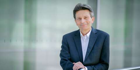 Rolf Mützenich steht vor einer Glaswand und lächelt in die Kamera.