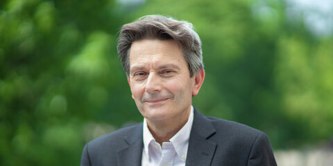 Rolf Mützenich steht auf einem Balkon mit grünen Bäumen im Hintergrund und schaut lächelnd in die Kamera.