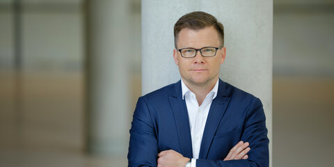 Carsten Schneider ist an einen Betonpfeiler angelehnt und blickt frontal in die Kamera