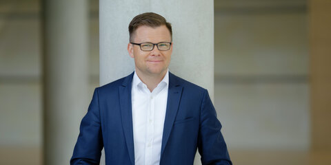 Carsten Schneider ist an einen Betonpfeiler angelehnt und blickt lächelnd in die Kamera.