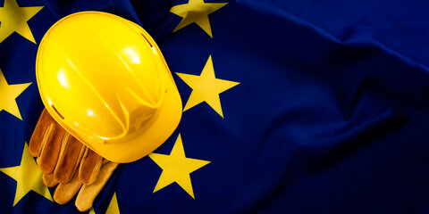 Ein gelber Schutzhelm und Schutzhandschuhe, im Hintergrund ist die EU-Flagge zu sehen