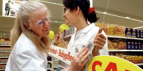 Zwei Frauen hängen in einem Supermarkt ein Schild auf. In dem Supermarkt arbeiten Menschen mit und ohne Behinderungen.