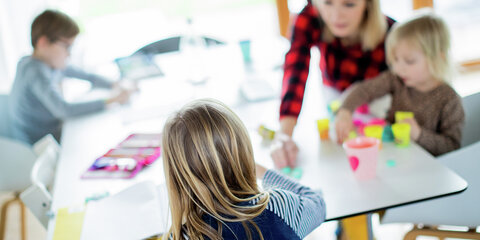 Eine Lehrerin sitzt mit ihren drei Kindern im Wohnzimmer, unterrichtet ihre Schüler einer Berufsschule im Distanzunterricht und hilft ihren Kindern bei ihren Aufgaben für die Schule.