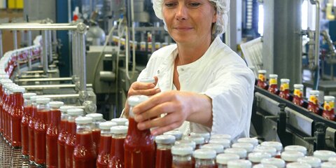 In einer Produktionshalle laufen Flaschen mit Ketchup auf einem Förderband und werden kontrolliert.
