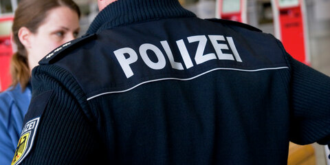 Rückansicht eines deutschen Polizeibeamten im Dienst, der eine junge Frau am Flughafen überprüft. Auf der Rückseite der Uniformjacke ist "POLIZEI" zu lesen.