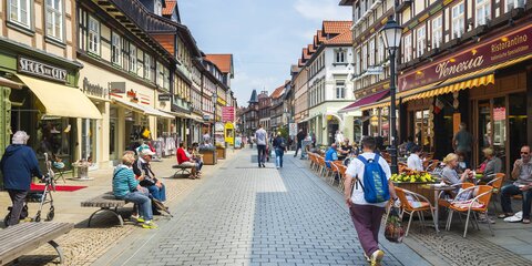 Blick in eine Fußgängerzone mit Geschäften in historischen Häusern. Menschen gehen die Straße entlang oder sitzen auf Stühlen in einem Café.