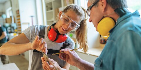 Tischlermeister und Auszubildende, die ein Holzstück in einer Werkstatt messen.