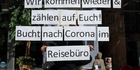 Ein Mitarbeiterin eines Reisebüros klebt einzelne Blätter mit der Aufschrift „Wir kommen wieder & zählen auf Euch! Bucht nach Corona im Reisebüro“ in das Schaufenster.