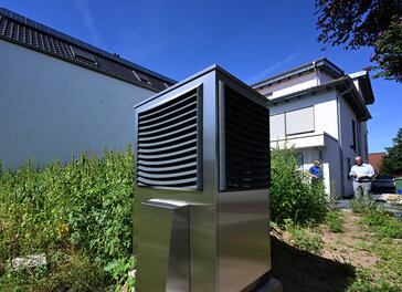 Vor einem neu gebauten Wohngebäude ist im Garten eine Wärmepumpe zum Heizen des Gebäudes in Betrieb.