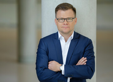 Carsten Schneider ist an einen Betonpfeiler angelehnt und blickt frontal in die Kamera